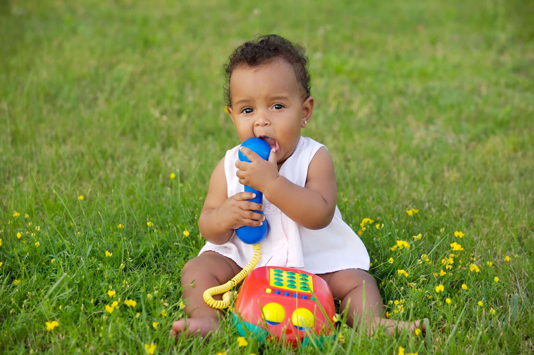 a baby sitting in grass teething on a plastic toy